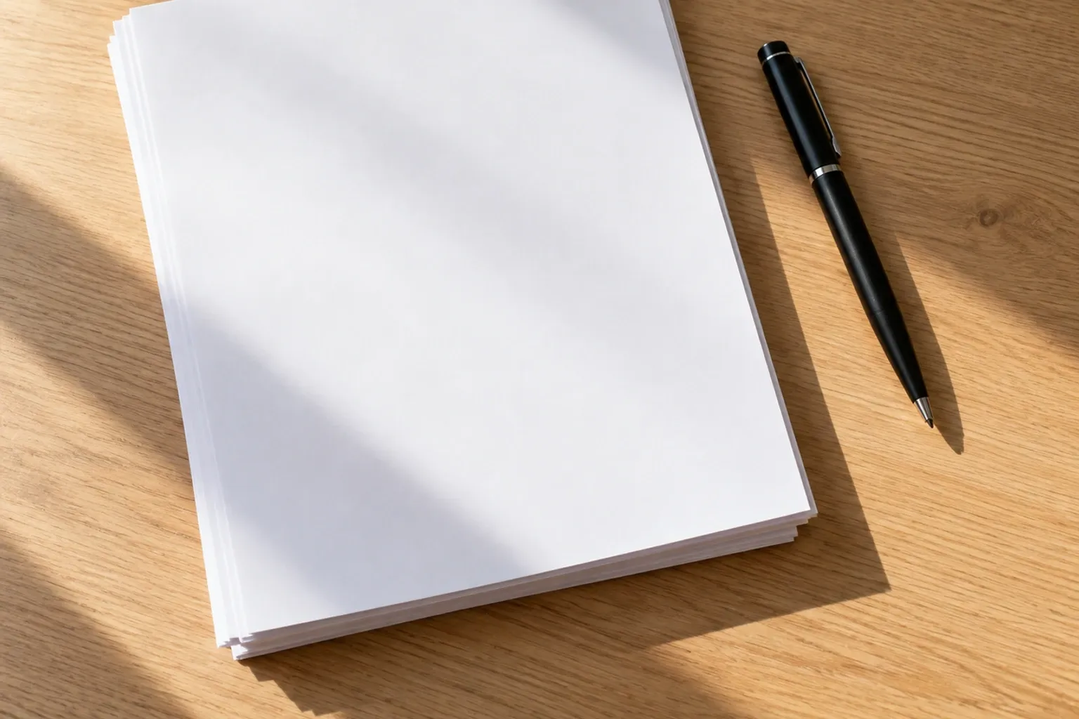 Stapeltje blanco papieren naast zwarte pen op houten bureau met zonlichtstrepen